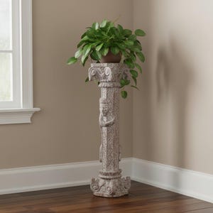 May include: A tall, light gray stone pedestal stand featuring intricate carved details, including a central figure and ornate top and bottom capitals. A lush green potted plant with rounded leaves sits securely on the flat top surface.