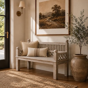 May include: A light-toned wooden bench with a spindle back and lattice side panels, featuring a square beige throw pillow and a rectangular beige lumbar pillow with a vertical brown stripe, positioned in a room with a framed landscape print.
