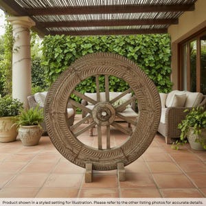 May include: A large, circular wooden wagon wheel featuring a carved, textured rim with repeating diagonal patterns. The wheel has a central hub and multiple spokes, supported by two small wooden blocks at the base on a tiled floor.