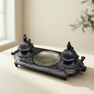 May include: An antique inkwell set with a dark, metallic finish. The set includes two ink pots with ornate lids and a central dish, all resting on a decorative tray. The design features intricate patterns and a vintage aesthetic.