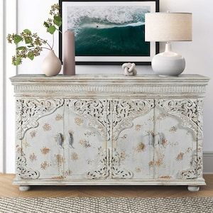 May include: A rectangular wooden sideboard featuring four doors with intricate carved scrollwork details. The finish is a distressed light gray paint with visible brown wood undertones. The cabinet stands on four small bun feet and includes metal hardware.