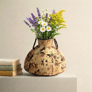May include: A round, weathered metal vase with two side handles, featuring a distressed beige and brown finish. The vase is filled with a bouquet of purple lavender, white daisies, and yellow goldenrod flowers, positioned next to three stacked books.