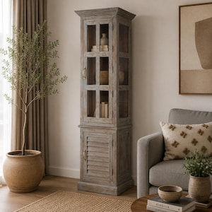 May include: A tall, narrow wooden cabinet with a weathered gray finish. The upper section features a glass-paneled door with a brass handle, revealing interior shelves. The lower section has a solid door with horizontal slats and a brass handle.