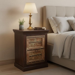 May include: A dark brown wooden nightstand featuring a single cabinet door with a distressed, weathered light-colored wood panel insert, metal trim, and a circular ring pull handle. The top surface holds a brass-toned lamp and a small potted plant.