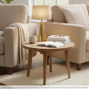 May include: A round wooden side table with three tapered legs, featuring a light-colored wood grain finish. On the table sits a gold-toned conical lamp with a beige shade, a small ceramic bowl, a stack of white books, and reading glasses.