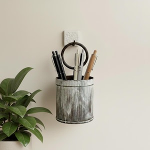 May include: A rustic, cylindrical pen holder made of distressed metal, mounted on a wall. The holder contains a black, silver, and brown pen. A decorative ring and floral tile are above the holder. A potted plant is in the lower left corner.