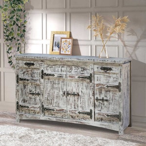 May include: A distressed white wooden sideboard with black hardware. The cabinet has multiple doors and drawers, with decorative metal hinges and latches. Two framed pictures and a vase with dried plants sit on top.
