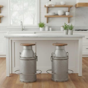 May include: Two galvanized metal bar stools with wooden seats. The stools have a milk can design with a footrest and handles. The stools are in a kitchen setting with a white island and wooden floor.