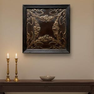 May include: A square, dark brown framed wall art with a gold floral design. Two gold candle holders with lit candles are on a dark brown table. A decorative bowl sits on the table.