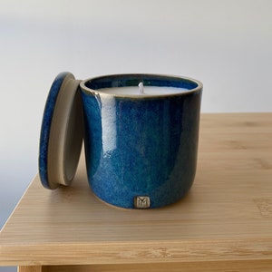 May include: A blue ceramic candle jar with a white lid and a white candle inside. The jar has a dark blue glaze and a subtle green hue. The candle jar has a small, white "M" on the front.