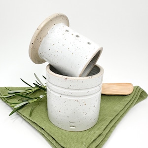 Puede incluir: Una mantequera de cerámica blanca con diseño moteado, con tapa y base con ranuras horizontales. El plato está sobre un paño verde con una ramita de romero y una pequeña espátula de madera.