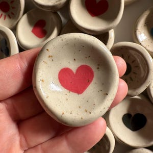 Peut inclure: Une collection de petites coupelles en céramique faites à la main. Chaque coupelle est beige avec une texture mouchetée et présente un motif peint à la main, notamment des cœurs rouges, des cœurs noirs et des soleils. Les coupelles sont rondes et semblent être de petits ramequins.