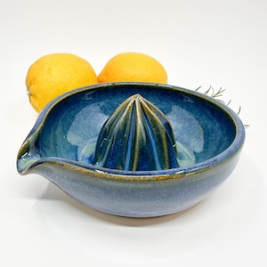 Op de afbeelding: Handgemaakte keramische citruspers in blauw met een tuit. Twee gele citroenen staan op de achtergrond. De pers heeft een geribbeld midden voor het persen van sap. Een stijlvol keukengereedschap voor vers sap.