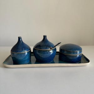 Peut inclure: Un ensemble de trois plats de service en céramique émaillée bleu foncé, chacun avec un couvercle, reposant sur un plateau rectangulaire. L'ensemble comprend deux pots couverts et un avec une petite cuillère. Le plateau a un bord blanc.