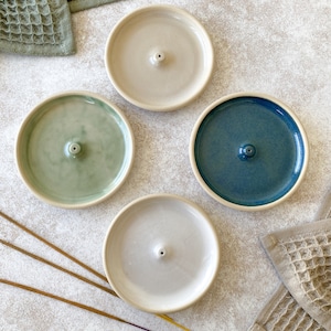 Può includere: Quattro porta incenso in ceramica nei toni crema, verde, blu e bianco. Ogni supporto ha un foro centrale per i bastoncini di incenso. Sono visibili anche bastoncini di incenso e due asciugamani a nido d'ape. I supporti sono disposti su una superficie chiara.