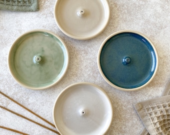 Könnte beinhalten: Vier Keramik-Räucherstäbchenhalter in Creme, Grün, Blau und Weiß. Jeder Halter hat ein zentrales Loch für Räucherstäbchen. Räucherstäbchen und zwei Waffelhandtücher sind ebenfalls sichtbar. Die Halter sind auf einer hellen Oberfläche angeordnet.