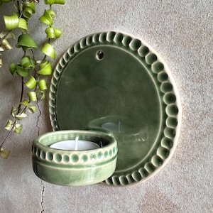May include: A green ceramic wall-mounted candle holder with a round plate and a small bowl. The plate has a decorative edge, and the bowl holds a white candle. The holder is mounted on a textured wall, with green plants in the background.
