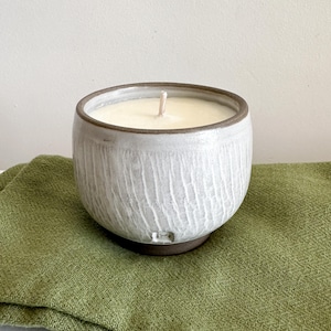 Op de afbeelding: Een handgemaakte keramische kaars in een ronde, komvormige vorm. De kaars is wit met een lichtbruine rand en een enkele lont. Hij staat op een groene doek, wat een minimalistische uitstraling creëert. De buitenkant van de kaars heeft een ruwe, gebeeldhouwde textuur.