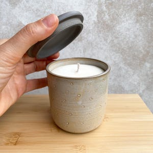 Peut inclure: Une bougie en céramique beige dans un récipient cylindrique tacheté avec un couvercle assorti. La bougie a une surface de cire blanche et une seule mèche. Le récipient est posé sur une surface en bois.