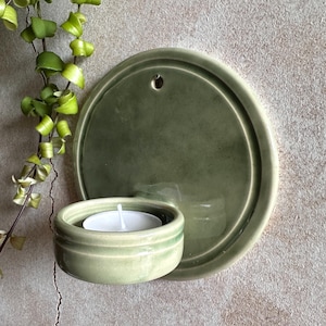 May include: A sage green ceramic wall-mounted candle holder with a round backplate and a small, round cup to hold a tea light candle. A white tea light candle is inside the cup. A trailing green plant is in the upper left corner.