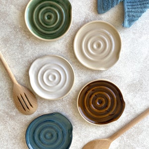 May include: Set of five ceramic spoon rests in various colors, including green, beige, brown, and blue. Each rest has a circular shape with a raised center and a curved edge for holding spoons.