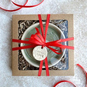 May include: A green ceramic bowl with a white rim is wrapped in a red ribbon and placed in a brown box with shredded paper. A small white ceramic tag with the text "When in doubt, love" is attached to the ribbon.