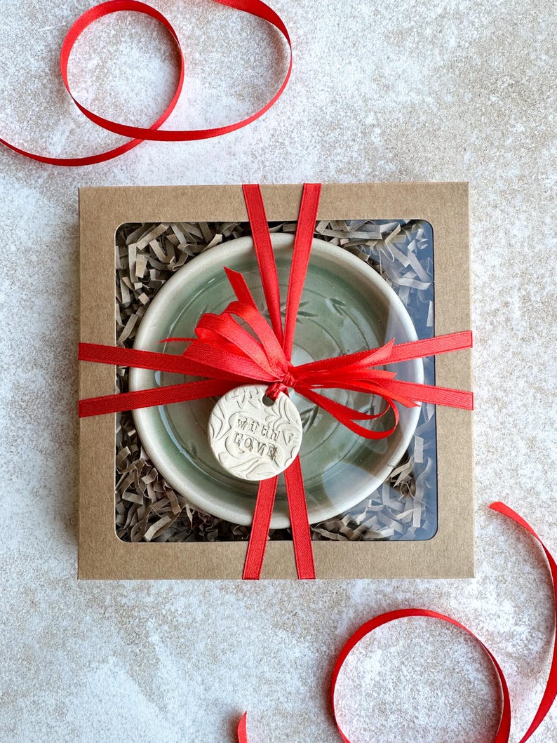 May include: A green ceramic bowl with a white rim is wrapped in a red ribbon and placed in a brown box with shredded paper. A small white ceramic tag with the text "When in doubt, love" is attached to the ribbon.