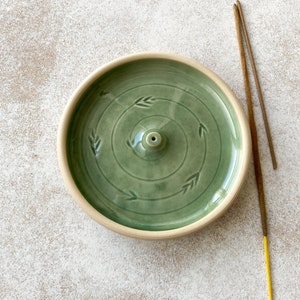 May include: A green ceramic incense burner with a circular design and a small hole in the center. Two incense sticks are shown next to the burner.