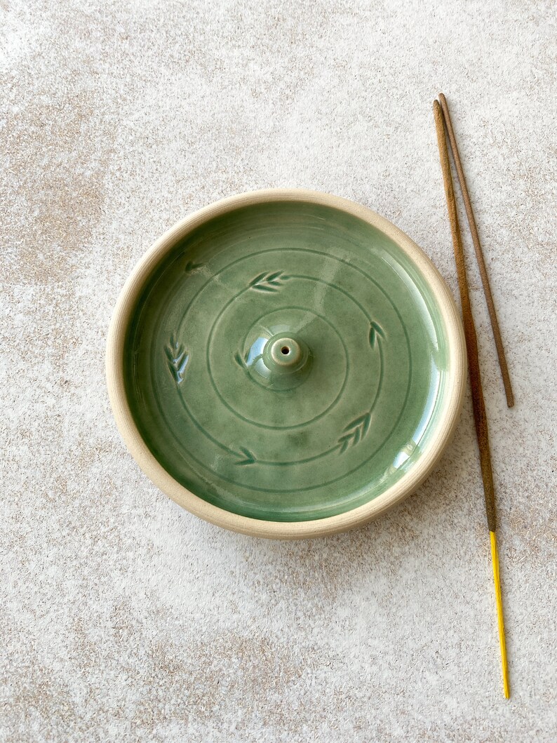 May include: A green ceramic incense burner with a circular design and a small hole in the center. Two incense sticks are shown next to the burner.