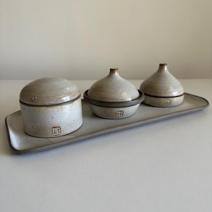 Pode incluir: Um conjunto de servir em cerâmica com uma bandeja retangular e três peças a condizer. O conjunto inclui um pote com tampa e dois recipientes com tampa, todos num esmalte bege salpicado com acabamento castanho escuro. Cada peça tem um pequeno logótipo quadrado.