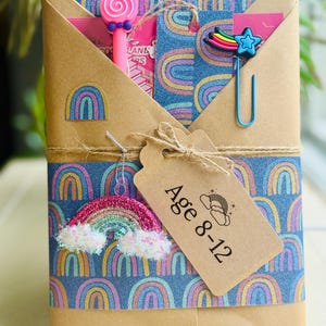 May include: A wrapped gift box with a rainbow theme. The box is wrapped in brown paper and blue paper with rainbow patterns. It is decorated with a rainbow charm, a star-shaped paper clip, and a tag that reads "Age 8-12".