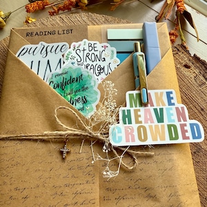 Puede incluir: Un sobre de papel kraft con una lista de lectura y pegatinas con las frases "pursue", "Be Strong and Courageous" y "Make Heaven Crowded". El sobre está atado con hilo y tiene un pequeño colgante de cruz.