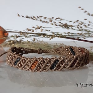 Peut inclure: Bracelet en macramé marron fait à la main, avec un motif tissé orné de petites perles bronze. Des touches de perles noires et orange ajoutent du détail. Le bracelet est réglable et convient à différentes tailles de poignet.