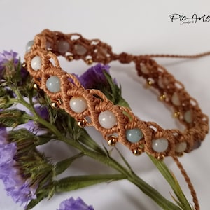 May include: A brown macrame bracelet with light green and white beads and gold accents. The bracelet is on a white background with purple flowers.