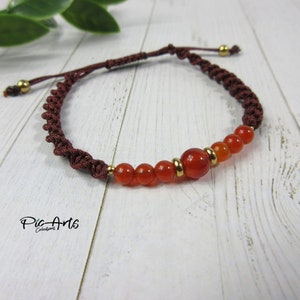 May include: A handcrafted bracelet with orange-red beads and a central bead, complemented by gold-coloured metal rings. The bracelet is made with a dark brown, braided cord and adjustable sliding knots. The bracelet is approximately 18 cm in length.