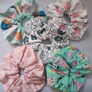 May include: A collection of five fabric scrunchies in various colors and patterns. The scrunchies feature prints of cartoon characters and floral designs in shades of pink, green, and white. These hair accessories are ideal for adding a touch of style.
