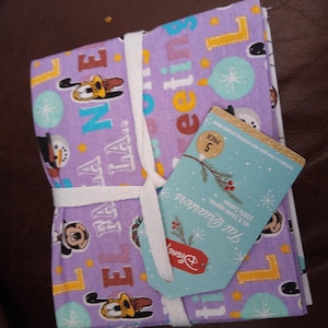 Peut inclure: Un lot de coupons de tissu Disney, attachés avec un ruban blanc. Le tissu est violet avec des personnages de dessins animés, des flocons de neige et les mots "Fa La La" et "Sweet". Une étiquette bleue avec une brindille et le logo Disney est attachée.