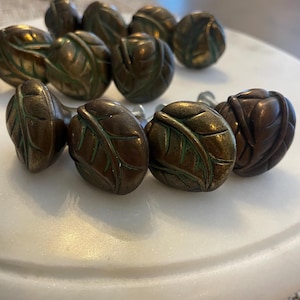 May include: A collection of round metal cabinet knobs featuring a raised leaf pattern. The knobs have a dark bronze finish with subtle green oxidation in the recessed areas, creating a textured appearance on each individual piece.