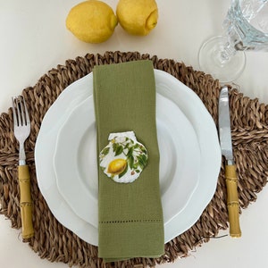 May include: A green linen napkin with a white seashell with a lemon design on it. The napkin is placed on a white plate with a brown woven placemat.