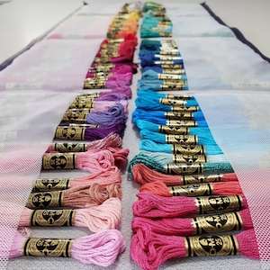 以下が含まれることがあります： 色別に並べられたDMC刺繍糸の糸巻きコレクション。糸巻きにはDMC番号と「Mouline Special」と「Mouline」の文字が金色の文字で書かれています。