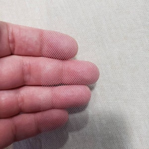 以下が含まれることがあります： 白いチュール生地のクローズアップで、繊細なメッシュ織りがわかります。生地は手で持たれており、柔らかく透き通った質感です。