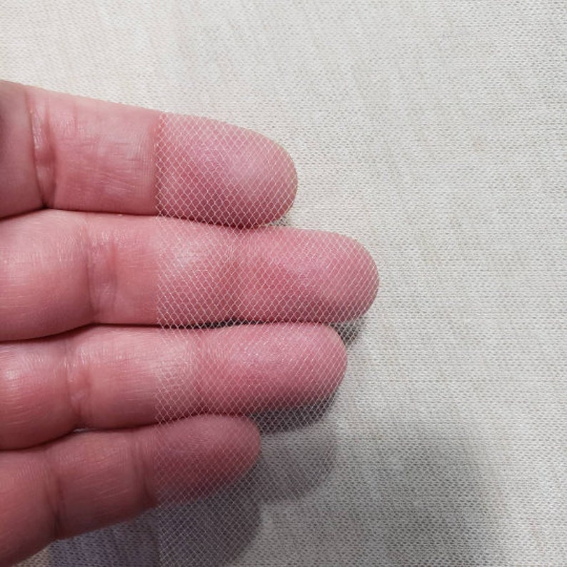 以下が含まれることがあります： 白いチュール生地のクローズアップで、繊細なメッシュ織りがわかります。生地は手で持たれており、柔らかく透き通った質感です。