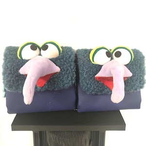 May include: Two square pouches featuring a character design with a textured dark teal fleece top flap, a smooth purple base, large white eyes with yellow and green borders, and a prominent, long, light purple nose above a red mouth opening.