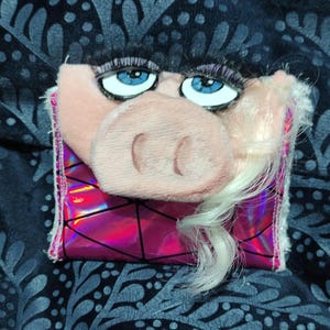May include: A plush wallet featuring a pig face with blue eyes, long black eyelashes, and blonde synthetic hair. The lower section is made of a shiny, metallic pink material with a black geometric grid pattern and white fuzzy trim on the sides.