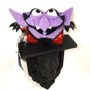 May include: A plush purple vampire character with large white eyes, black eyebrows, and a black goatee sits atop a red metallic box. This box rests on a black rectangular shelf mounted above a black wall plaque featuring a sculpted face.