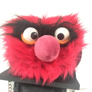 May include: A plush, bright red, fuzzy character bag featuring large white eyes with black pupils, thick black eyebrows, and a prominent round pink nose. The bag is attached to a black, rectangular, structured base with a silver-toned metal chain strap.