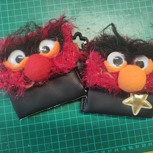 May include: Two small pouches featuring fuzzy red and black textured tops with large googly eyes and round noses. The bottom sections are made of smooth black material. One pouch includes a gold star applique, and a red star-shaped charm is attached.