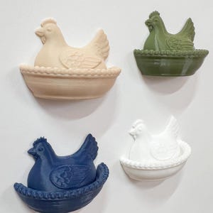 Könnte beinhalten: Vier dekorative Hühnerfiguren im Nestdesign. Die Hühner sind in Beige, Grün, Marineblau und Weiß gehalten. Jedes Huhn sitzt in einem farblich passenden Nest. Die Figuren sind aus einem festen Material gefertigt.