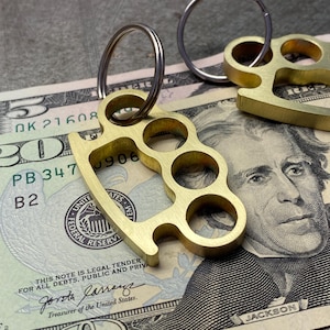 May include: A brass key chain with a key ring and a brass knuckle design. The key chain is lying on a twenty dollar bill with the text "OK 21608", "PB 347", "B2", "UNITED STATES", "FEDERAL RESERVE", "THIS NOTE IS LEGAL TENDER FOR ALL DEBTS, PUBLIC AND PRIVATE", "Jorotha Jarrany", "Treasurer of the United States", and "JACKSON".