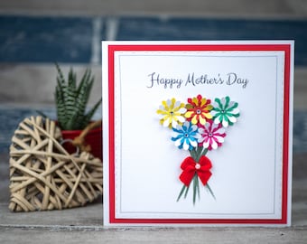 May include: A handmade Mother's Day card with a bouquet of colorful paper flowers. The card has a red border and the words "Happy Mother's Day" in script. A small red bow adorns the flower stems. A heart-shaped woven decoration and a potted plant are in the background.